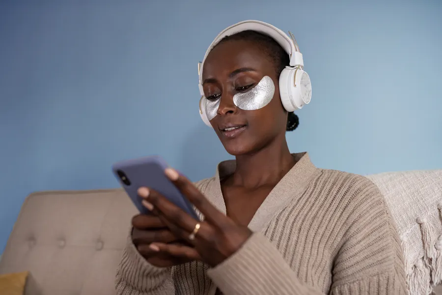 Woman wearing under eye mask wearing headphones and browsing onphone